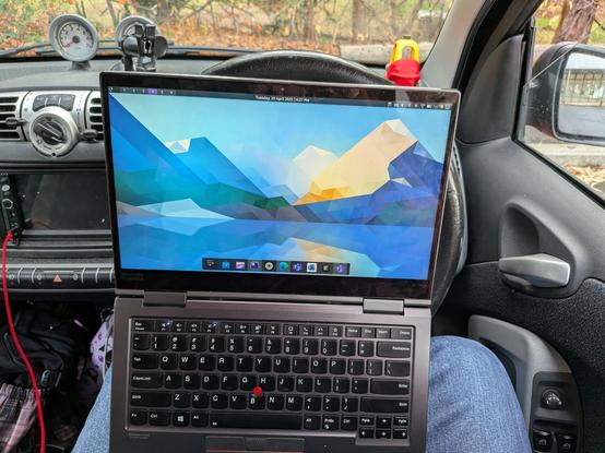 A Lenovo yoga laptop mostly open leaning on the steering wheel of a car. 

The KDE desktop wallpaper is mostly blue and you can see a Little Tykes model car on the dashboard. 

The car is parked in front of some trees on an overcast day.