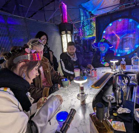 4 people sitting at a marble bar and one standing behind. They are playing a dice game. Outside the bar is a colorful background.
