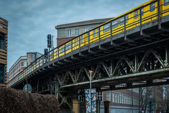 Ein erhöhter gelber Zug fährt über eine Stahl-Fachwerkbrücke, vor dem Hintergrund städtischer Gebäude unter einem bewölkten Himmel. Eine Stange mit Aufklebern und ein Verkehrszeichen sind im Vordergrund zu sehen.