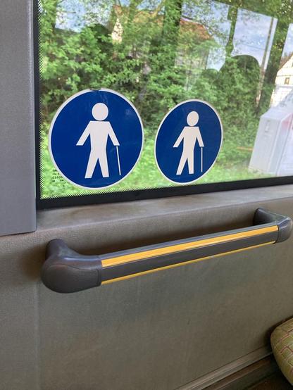 Zu sehen ist eine Busfensterscheibe aus dem Businneren fotografiert. Darauf machen zwei nebeneinander angebrachte Aufkleber mit weißen Figuren mit Gehstock auf blauem Hintergrund darauf aufmerksam, dass die dazu gemeinten Sitzplätzen für alte Menschen und Schwerbehinderte Personen freigemacht werden sollen (müssen).