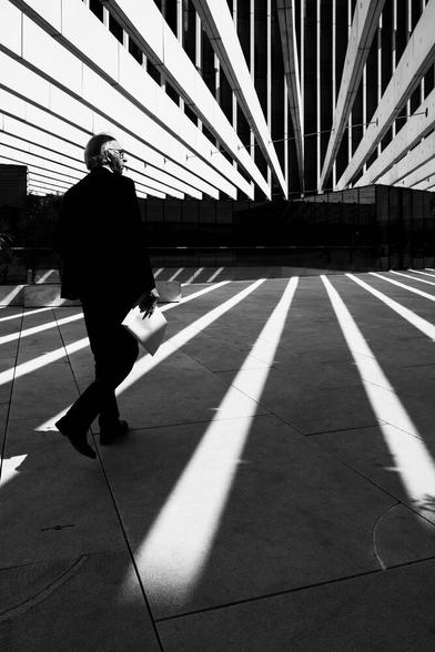 A man in a suit holds a big envelope. He walks towards a black and white building. There are strips of light on the ground, and white panels above it, forming leading lines for the subject.