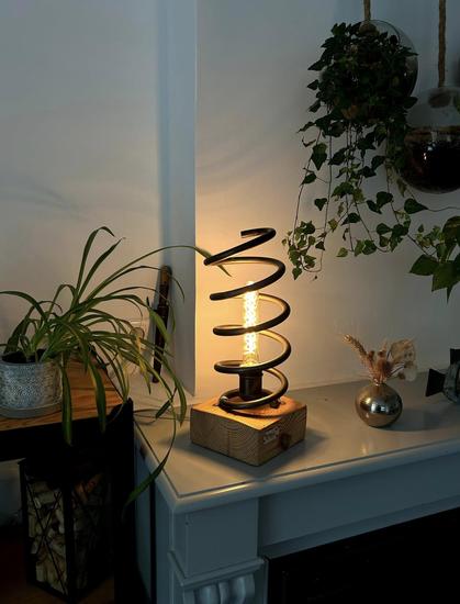 Lampe constituée d'un socle de bois clair, sur lequel est fixé debout un amortisseur de Honda Civic, avec en son sein une ampoule à filament, allumée. La lampe est posée sur un coin de cheminée, entourée de plantes vertes