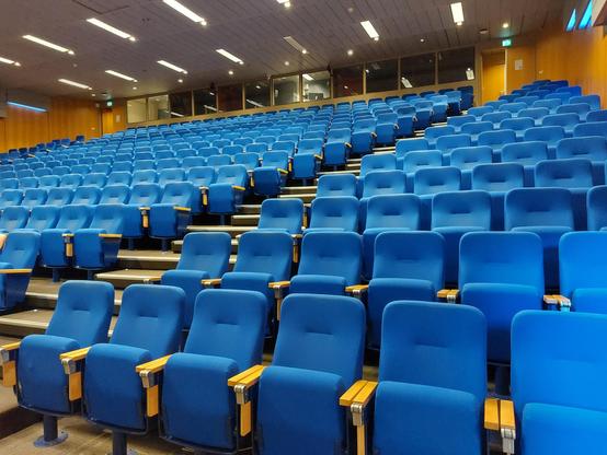 Lecture hall with empty blue upholstered seats in rows, wooden arms, and a softly lit ceiling.