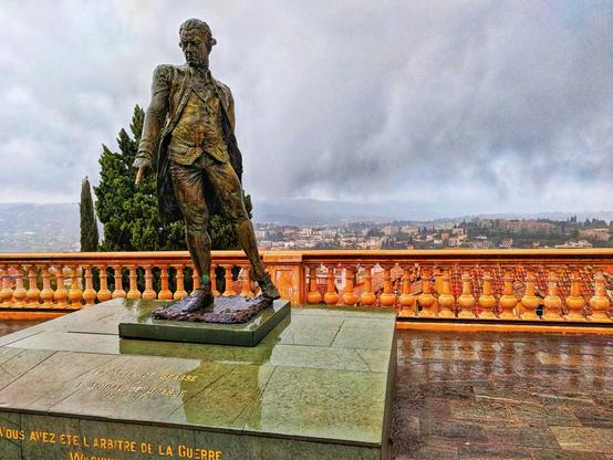 A photograph shows a bronze statue of Admiral de Grasse, standing on a plinth. The statue is depicted in a formal pose, with the Admiral wearing a period military uniform. The statue is positioned outdoors, with a decorative orange balustrade behind it and a cityscape in the background under a cloudy sky. The plinth is inscribed with text, including the name "AMIRAL DE GRASSE". The photo is taken at a low angle, emphasizing the statue's height and presence.