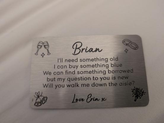 The image shows a metallic card with a brushed finish, placed on a light-colored surface. The card features a message engraved on it, addressed to "Brian." The text reads:

"Brian

I'll need something old

I can buy something blue

We can find something borrowed

but my question to you is new

Will you walk me down the aisle?

Love Erin x"

The card is decorated with small illustrations: two clinking glasses at the top left, two interlocking rings at the top right, two bells at the bottom left, and a bouquet of flowers at the bottom right. The text is in a mix of cursive and print fonts, with the name "Brian" and the signature "Love Erin x" in cursive. The overall design is elegant and romantic, suitable for a wedding proposal