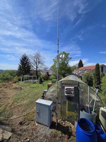 Foto von einem grauen-Kabelverzweiger KVZ59, daneben ein 4m Antennenmast mit 2 Outdoor-APs, einer Richtfunk-Antenne, einer kurzen Antenne darüber eine längere Antenne mit 3 Radials. rechts blaue Regenwassertonnen. Im Hintergrund: ein kleines Gewächshaus, grüne Wiese mit teils aufgewühlter Erde, Reihehaus-Reihe, Horizot, blauer Himmel mit weißen Cirrus-Wölkchen.