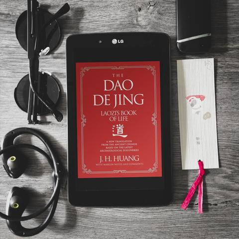 the dao de jing laozi's book of life translated by j.h. huang, red cover of a new translaton on taoism father's philosophy book. black-framed LG tablet is placed on a dark grey wooden table, with various dark items strewn around, such as round black shades, earphones, bookmark, lighter. review by reading nosferatu, image by vakaris aka wannabekingpin