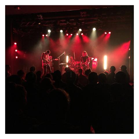 Quadratisches Foto. Eine dreiköpfige Band auf einer Bühne, von den Scheinwerfern werden die Musiker rot angestrahlt. Davor die Silhouetten tanzender Menschen.