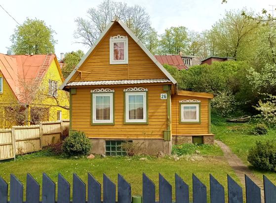 A yellow log house in typical regional style with horizontal exterior planking. Thereis a basement window in the plastered foundation. The windows are white PVC with exterior hinges and inserted plastic cross to emulate the look of the original smaller window panes. There is a green painted wooden trim and a white painted elaborate cutout at the top. The corner trim is painted matching green as well.