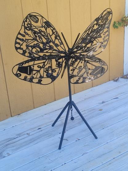 Sur la photo une sculpture de jardinbennforme de papillon en acier peint en noir. 

Les ailes du papillon sont faites de toutes sortes de rebut d'acier.  Il est déposé à l'extérieur sur une terasse blanche devant une maison jaune.