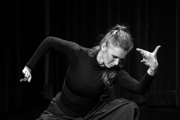 Black and white photo of a dancer. She is dressed in black and photographed from the front. She bends forward and to the left, left arm bent upwards, hand gracefully bent. Right arm bent downwards. Her face is the most striking, in the light, very concentrated, her long blond hair tied in a ponytail, falls over her left shoulder.