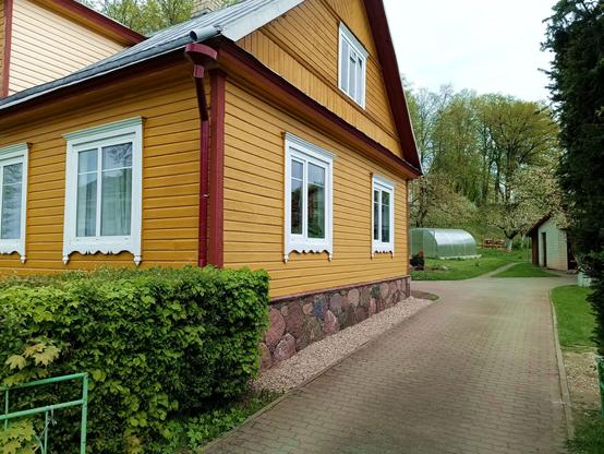 Another yellow house with brown accents behind a hedge. The foundation is stones that appear to be lose but probably are cemented in from the back, as they are too perfectly aligned to be original. The window siding is white with an elaborate bottom. The windows themselves are PVC split vertically into two panes. Some of them with one pane opening inwards. All panes have a horizontal plastic bar inserted into the glass package near the top, emulating the original split there. The attic window is wood, three vertical panes with a small openable section at the center pane top.

The roof is fold-joined metal and the gutter features an odd funnel near the top from a retrofitting downpipe. A new looking plastic greenhouse lurks in the garden behind. The driveway is recently paved cement stones.