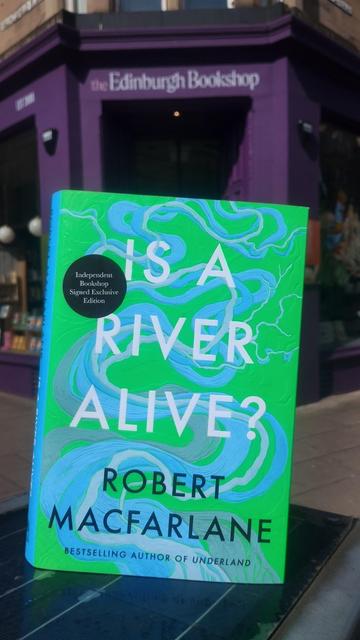 Green cover, criss-crossed by meandering, curving lines in shades of blue to denote rivers crossing the landscape, on the hardback of Is a River Alive? by Robert MacFarlane, outside the Edinburgh Bookshop. Cover painting by Stanley Donwood