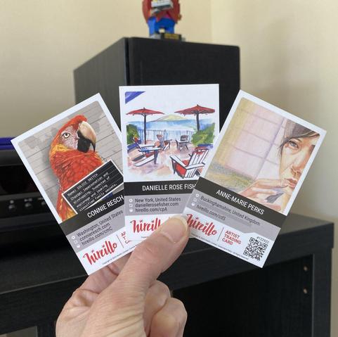 A hand holding three artist business cards featuring artwork: a parrot, a patio scene, and a portrait.