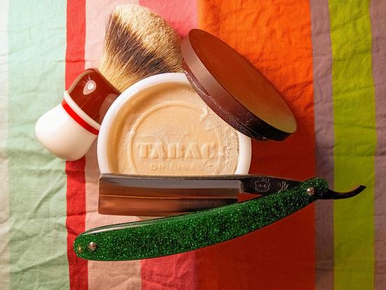 (Le Chat Mistral description)

This image shows a classic shaving set arranged on a colourful striped cloth. There's a shaving brush with a dark handle and light-coloured bristles, placed in a white and red mug. N.ext to it is a round shaving soap in a white bowl with "Tabac" written on it. A straight razor with a shiny green handle is laid across the bowl. The razor's blade is open and has a dark handle. The cloth underneath has vertical stripes in various colours, including green, orange, and red. The items are neatly organised, giving a vintage and elegant feel to the setup.