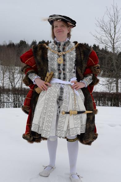 A person dressed in an elaborate Tudor-style historical costume stands outdoors in a snowy landscape. Resembling Henry 8th. The outfit includes a richly decorated doublet, a fur-trimmed red cloak, a patterned white skirt, white tights, and flat shoes. They are adorned with gold jewelry, including a large medallion necklace and multiple rings, and wear a black hat with a feather. Trees and snow-covered ground are visible in the background.