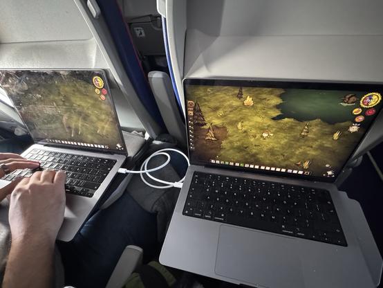 A photograph of two MacBooks placed on airplane tray tables, connected by a Thunderbolt cable and playing a LAN game of Don't Starve Together.
