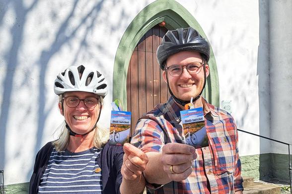 Zwei Personen stehen vor einem Gebäude mit einem markanten, grün umrandeten, gotischen Torbogen. Beide tragen Helme, was auf eine Aktivität wie Radfahren hinweist. Die Person links trägt einen weißen Radhelm und ein gestreiftes T-Shirt, während die Person rechts einen schwarzen Radhelm und ein kariertes Hemd trägt. Beide halten kleine, blaue Karten mit dem Text "Fahrradsegen" und "Bleib behütet" in der Hand. Die Umgebung ist sonnig, und die Schatten der Bäume fallen auf die Wand.

[by AltBot]