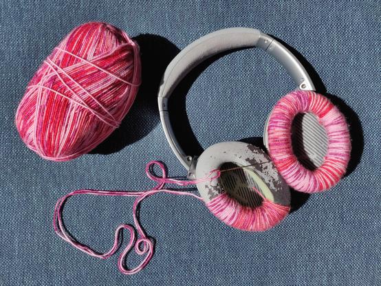 A pair of grey noise cancelling headphones with very worn out foam pads. The foam is peeping out of the split and peeling faux leather. One side has been repaired by wrapping it in pink and red yarn. The other side is a third of the way done, a trail of yarn still attached to a needle. The ball of yarn is pictured next to the headphones.