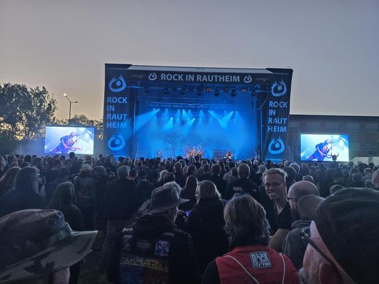 Foto vom Publikum beim Inklusions-Rock/Metal-Festival “Rock in Rautheim”. Es zeigt eine Mange Personen, die vor der Bühne stehne, auf der eine Rock-Band aufspielt. Die Bühne ist umgeben von Bannern mit dem Schriftzug “Rock in Rautheim”, die Außerdem das Logo der Lebenshilfe Braunschweig zeigen. Es besteht aus einem Punkt, der den Kopf darstellt und von einem oben leicht geöffneten Kreis ist, dessen Enden jeweils den typischen Metalgruß - die sogenannte Pommesgabel - bilden. So wirkt das Logo wie der stilisierte Oberkörper einer Person, die während eines mitreißenden Konzerts die Arme begeistert in die Höhe streckt, um de Band zu feiern. Links und rechts neben der Bühne steht jeweils eine große Leinwand, die das stattfindende Konzert noch besser für Personen in den hinteren Reihen darstellen. In diesem Fall zeigen sie einen singenden Gitarristen.