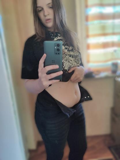 Foto von Lili vor dem Spiegel. Ungeschminkt und sichtbar angeschlagen. Sie trägt eine schwarze Highwaist Jeans die ihre Hüften gut betont, hat hier die Hose aber auf damit man ihren leicht speckigen Bauch sieht
