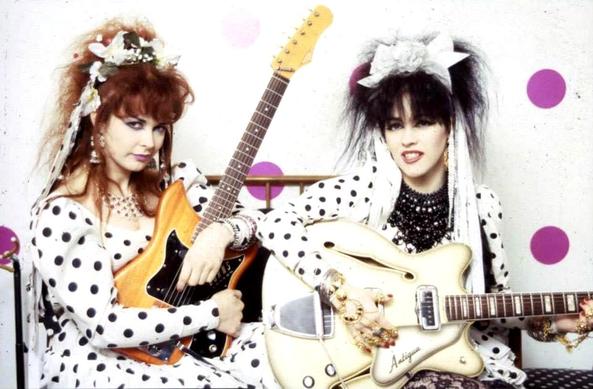 Strabwerry Switchblade - Jill Bryson on the left and Rose McDowall on the right, both in white polka dot dresses with white plastic flowers in their hair trailing polka dot ribbons. Each holding a guitar, and both looking at the camera.