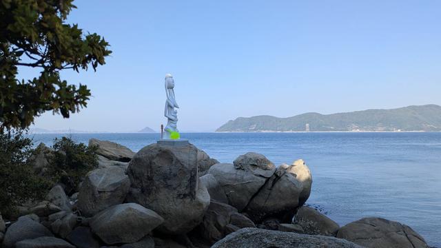 A Malian statue representing a mother and her child installed on a rock on the coast of the Seto Inland Sea of Japan. Instead of being made of wood, it's made of plastic (probably) and it's white and bright green instead of brown or black.