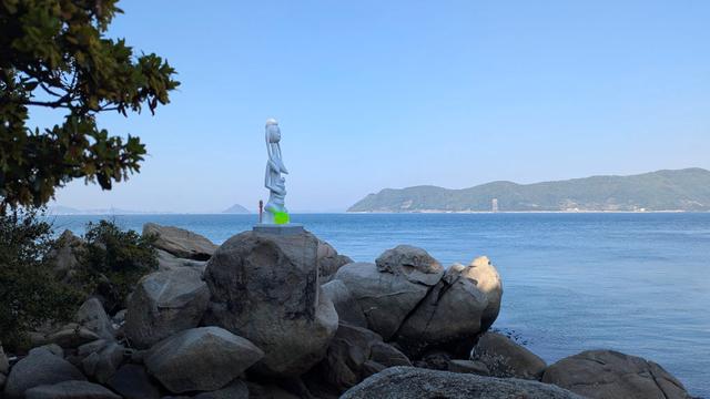 A Malian statue representing a mother and her child installed on a rock on the coast of the Seto Inland Sea of Japan. Instead of being made of wood, it's made of plastic (probably) and it's white and bright green instead of brown or black.