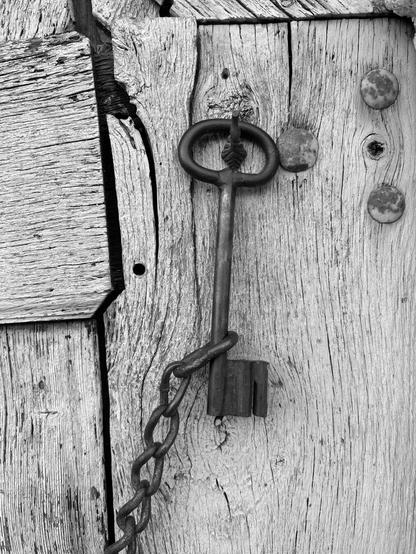This photo shows a close-up of a large, old metal key attached to a chain. The background is a wooden door.  The image is in black.