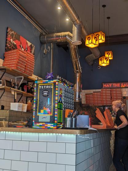The counter inside a pizza restaurant. Hanging light fixtures that look like coin blocks from Super Mario video games. Equipment on the counter encased in Lego blocks with action figures on top. A Spiderman print on the wall. More nerd stuff on shelves.