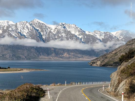 Scenic view of a large lake, taken through the windscreen of a tour bus driving down the road. Directly in front is the road, which then disappears to the right behind the foot of a mountain. In front of the road is a large blue lake, bordered in the distance by a range of snow-capped mountains. In the middle distance, floating above the lake, is a bank of very low cloud, almost at our eye-level.