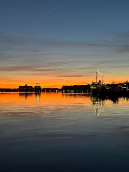 The glow of the sunset over water. There are the silhouettes and lights of buildings on the horizon, and a stationary ship with strings of lights on its masts closer to the foreground.