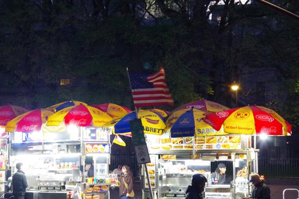 An American flag waves above a bike path sign between two halal carts at dusk.