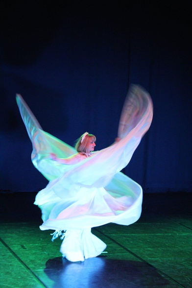 Tänzerin mit großem Seitenstoffkostüm an Stäben geführt in drehender Bewegung. Der Bühnenhintergrund ist blau und der Boden grün beleuchtet.