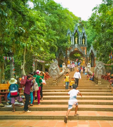 Photo shows old wide stone stairs up to a gate with statues from Hindu and Buddhist religion. The stairs are lightly crowded with people walking up and down, and some selling small items from small pop-up stores. In the centre, a young local boy is running up the stairs with several steps at the time. Two Garuda-guardian status keeps a Naga-snake statue each in front of the elaborate gates. It is a slightly overcast day, but the contrast between the stone stairs and statues, and the deep green of the surrounding forest.