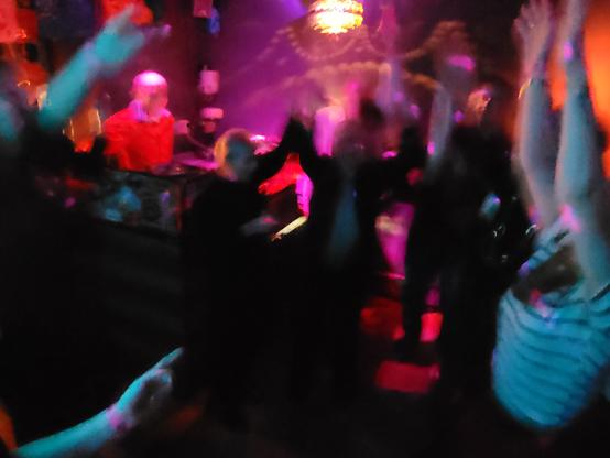 Querformat in Farbe. Ein unscharfes Bild einer kleinen Tanzfläche in rotpinkem Licht. Die tanzenden Menschen sind nur schemenhaft zu erkennen. Sie werfen ihre Arme nach oben. Nur der rot angeleuchtete DJ links im bild, steht still. Über der Tanzfläche dreht sich eine discokugel