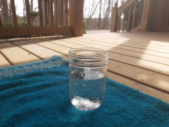 Photo near ground level of the corner of my blue blanket with a mason jar mostly full of water. The deck boards, rails, and a gap at the stairs opening up to bare branched trees in the distance. Feeling very chill and grounded. Hope sharing the image also shares the vibe.