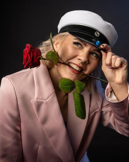 The image shows a person wearing a white cap with a black visor and a pink blazer. They are holding a red rose in their mouth and are adjusting the cap with their hand while smiling.