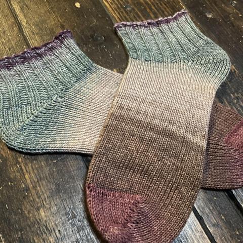 A pair of hand knit socks in shades of brown, taupe, and muted slate blue are laid flat on a wooden surface.