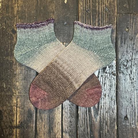 A pair of hand knit socks in shades of brown, taupe, and muted slate blue are laid flat on a wooden surface.
