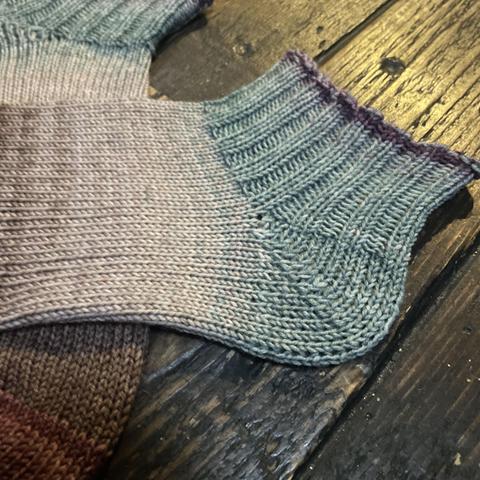 Detail of a pair of hand knit socks in shades of brown, taupe, and muted slate blue are laid flat on a wooden surface.