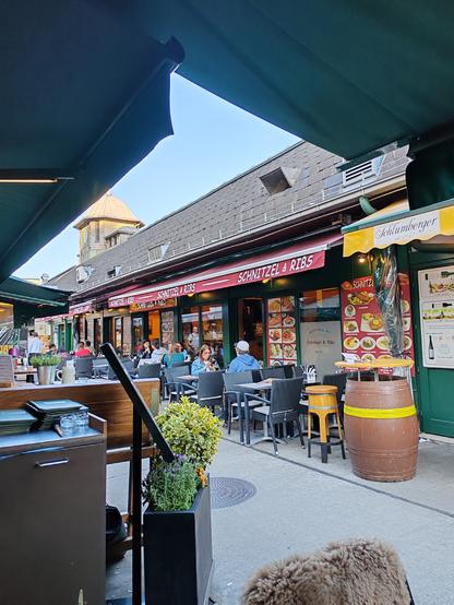 Picture taken from a table outside a bistro looking across an alley to an opposite located bistro selling schnitzel, ribs and other Viennese food. To the right are signs indicating the types of wine sold at a neighbouring wine bar. The blue sky is visible through a break in the overhanging rain canopy, offering shade.