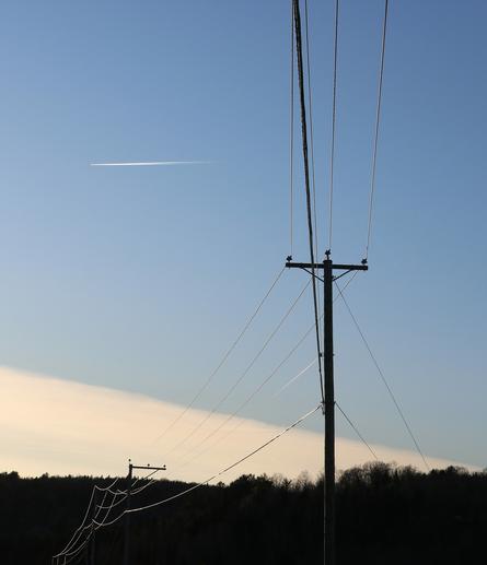 Photo of a small power line taken from under, its five different cables shining slightly in the fading light, and the pole only a dark silhouette against a blue sky. A plane with its white trail is flying in the sky in top left corner. The dark silhouette of the forest is seen in the distance and a white cloud with a triangular shape creates a diagonal line above the horizon.