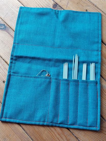 On a wooden floor is a pouch for double pointed needles I made. It is made from a thick teal fabric (although in the photo it came out way more blue/turquoise), with red stitching that creates several slots for the needles to sit in, with an extra wide slot for stitch markers, holders etc.