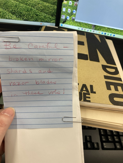 Handwritten note on lined paper: Be careful- broken mirror shards and razor blades in these vols. Paperbound volume in background.