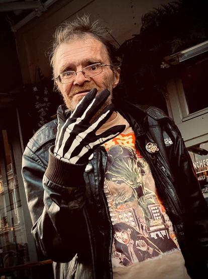 Portrait photographique d'Y5/P5 : un homme âgé, hirsute et avec les traits creusés. Il porte un blouson en cuir, des lunettes rectangulaire et un t-shirt illustré d'un monstre japonais. Sa main, au premier plan, est recouverte d'un gant imprimé d'une main de squelette. Il sourit.