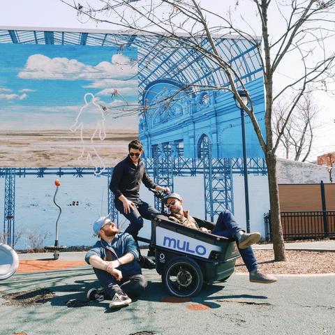 Three people siting on and around a cargo-bike in a park with a mural in the background. / Trois personnes assises autour et sur un vélo-cargo dans un parc, avec une murale en arrière-plan.