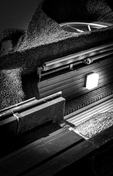 This black-and-white photograph shows the side of a building, shot from a low angle looking upwards. The roof is made of thatch, the structure of which is clearly visible. A rectangular light fixture is attached to the building's wooden cladding. Above the light fixture, a rolled-up awning is suspended below the eaves. A curved, white-framed window, integrated into the thatched roof, is partially visible at the top of the image. The slightly tilted perspective lends the composition an abstract and dramatic feel.