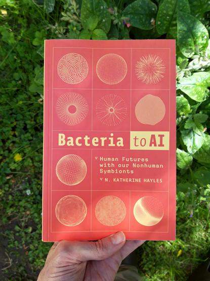 A hand holding a copy of the book "Bacteria to AI, Human Futures with our Nonhuman Symbionts" by N. Katherine Hayles.