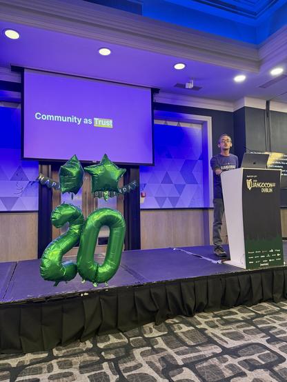 Me on stage at DjangoCon Europe. Slide says Community as **Trust**. There are "20" balloons for Django's birthday and I've got a DjangoNauts t-shirt on. The podium has the logo for the conference.